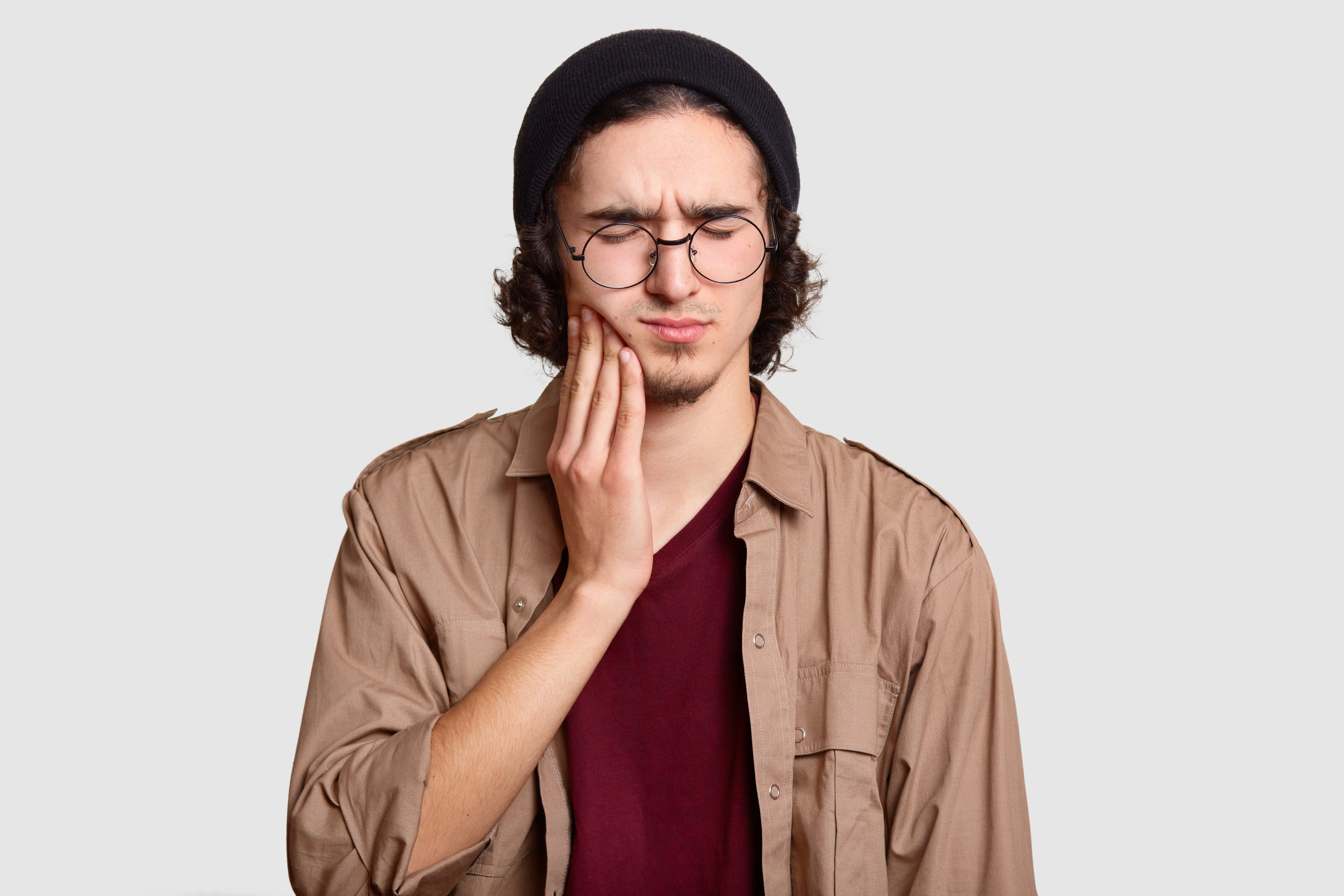 stressful youngster with small beard keeps hand cheek suffers from toothache keeps eyes shut dressed stylish clothes big round spectacles models white wall